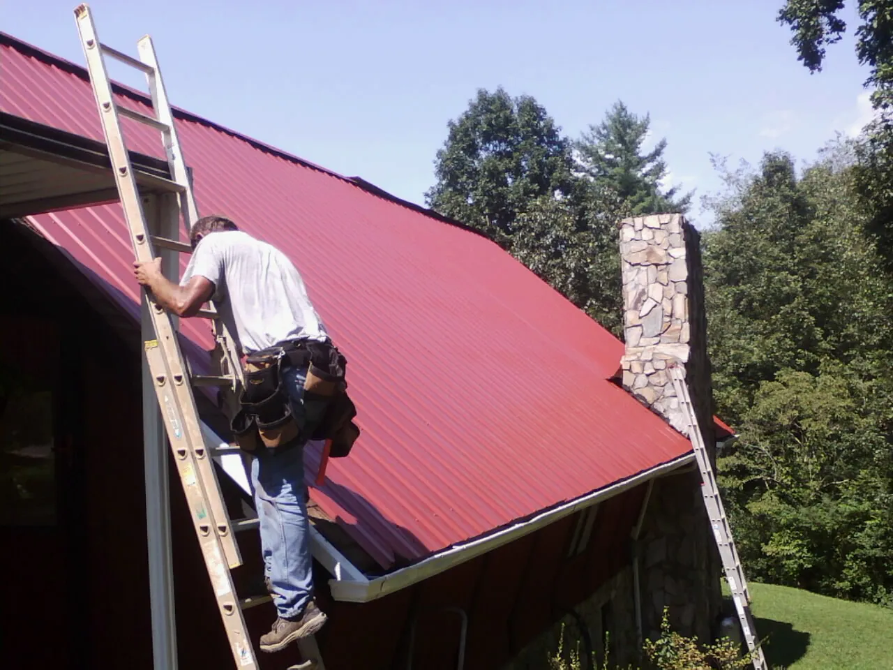 Roofer on a ladder beside a red metal roof and stone chimney — Soffit & Fascia Repair by Roofing Brothers in Frazee